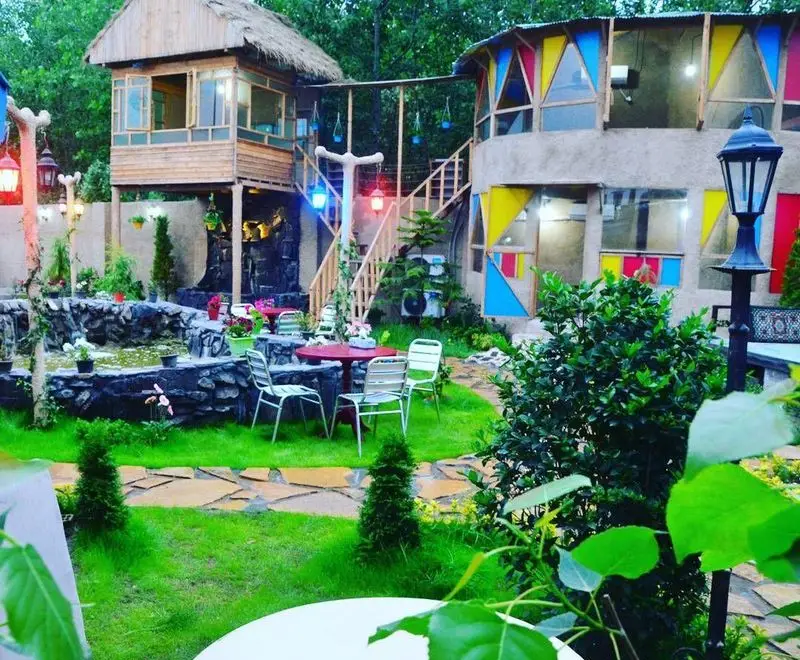 نمای دلنشین و زیبای رستوران گل یخ با کلبه چوبی منحصر به فرد، ساختمان رنگی، فضای سبز، آبنما و میز و صندلی در فضای باز.