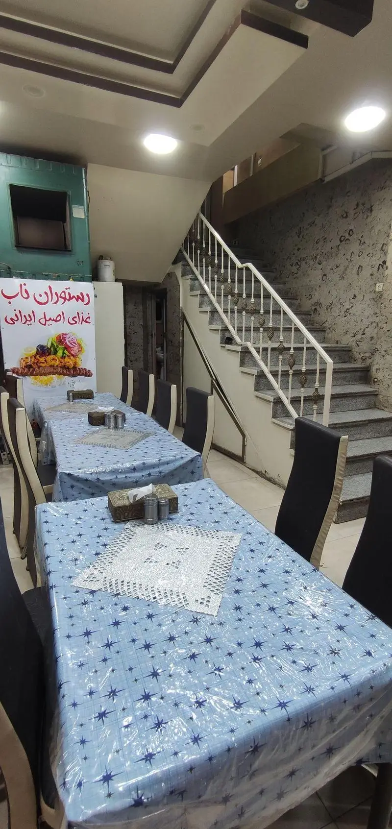 فضای داخلی رستوران ناب با میزهای چوبی پوشیده شده با رومیزی آبی ستاره‌دار، صندلی‌های شیک، تابلوی کباب ایرانی و پلکان سنگی.