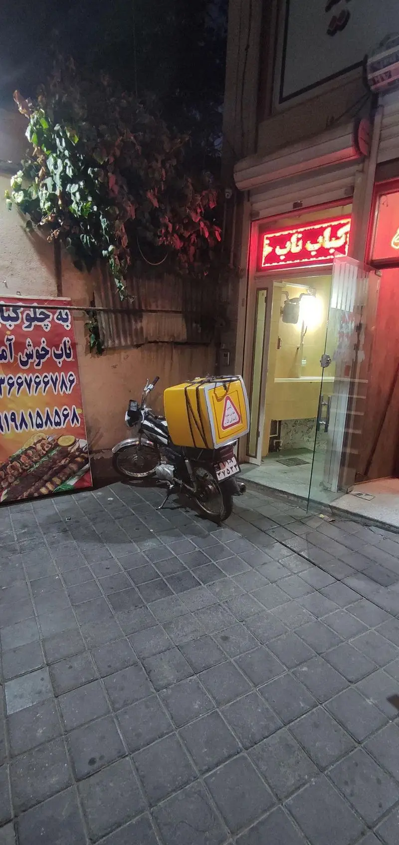 نمای شبانه رستوران کباب ناب با تابلوی نئون روشن و موتور دلیوری زرد رنگ، آماده سرو چلوکباب تازه و غذای ایرانی.