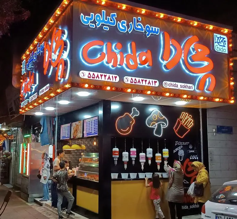 کسب و کار فست فود "سوخاری چی دا" با تابلوی نئونی جذاب و مشتریان در حال خرید مرغ سوخاری و سیب زمینی سرخ کرده، نمای شب.