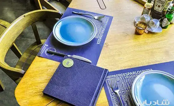 میز رستوران ایتالیایی اسپرلوس با بشقاب آبی و رومیزی شامل منوی پاستا و پیتزا. چیدمان آماده برای مشتریان.