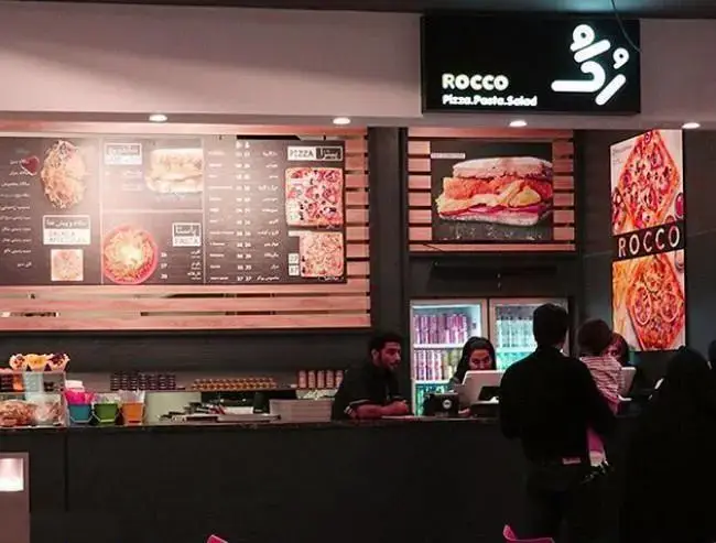 نمای داخلی رستوران فست فود راکو در مجتمع تجاری ارگ تهران، با منوی پیتزا، پاستا، سالاد و کارکنان در حال ارائه خدمات به مشتریان.