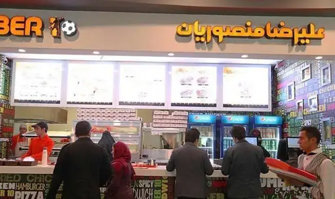 نمای داخلی فست فود شلوغ علی رضا منصوریان در مجتمع تجاری ارگ، مشتریان مشغول سفارش غذا و پرسنل در حال خدمت‌رسانی.