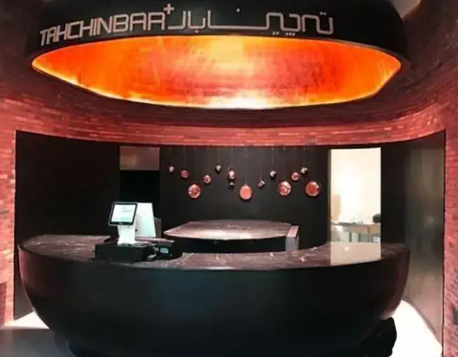 تصویری از طراحی داخلی شیک و مدرن رستوران تهچین بار در مجتمع تجاری ارگ شامل پیشخوان منحنی سیاه و سقف نارنجی نورانی و دکوری مسی.