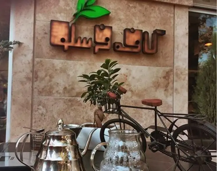 نمای بیرونی کافه فرسکو با تابلوی چوبی، لوگوی برگ سبز، کتری و قوری و دوچرخه تزئینی؛ فضایی دلنشین برای صرف قهوه و چای.