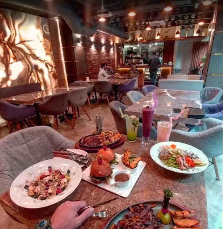 فضای داخلی کافه فرسکو با میز پر از غذاهای لذیذ شامل سالاد، برگر، استیک و نوشیدنی‌های خنک. محیطی شیک و دنج برای مشتریان.