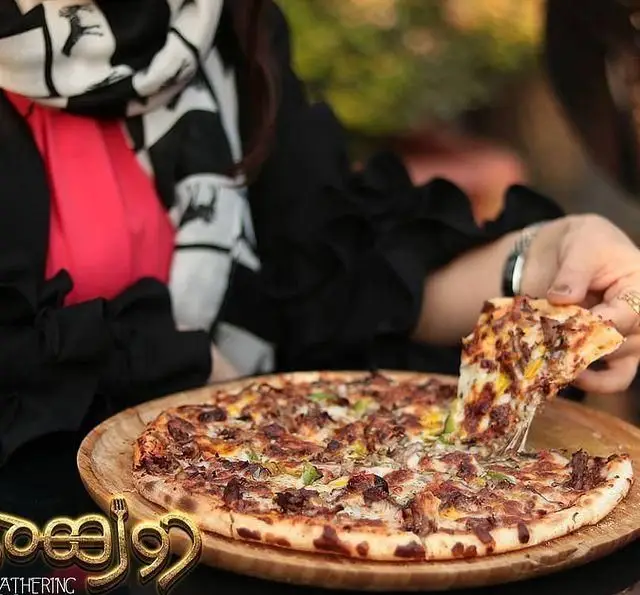 مشتری در حال برداشتن یک تکه پیتزای گوشت و پنیر کشدار از سینی چوبی در رستوران و کافی شاپ دورهمی، تجربه غذایی لذت‌بخش.