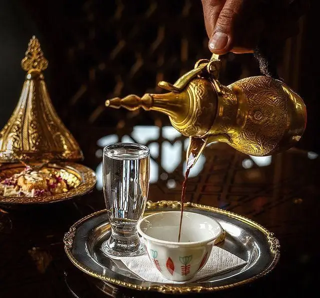 تصویری دلنشین از لحظه سرو قهوه عربی اصیل از دله سنتی طلایی همراه با لیوان آب و دسر شیک در رستوران و کافی شاپ دورهمی.