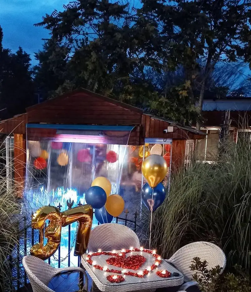 رستوران و کافی شاپ دورهمی: چیدمان رمانتیک فضای باز برای مناسبت خاص، شامل میز شمع و گل، بالون‌های 37 و آبنمای نورپردازی شده.