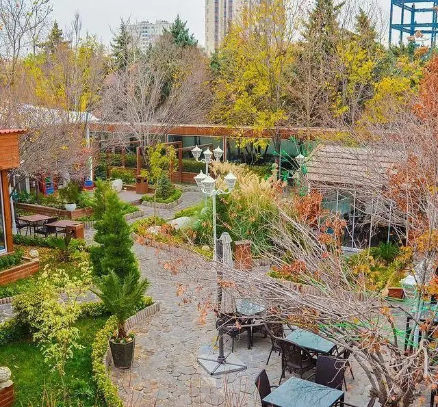 فضای باز زیبا و دنج رستوران و کافی شاپ دورهمی با میز و صندلی در میان باغ پاییزی پر از درختان، گیاهان و مسیر سنگفرش.