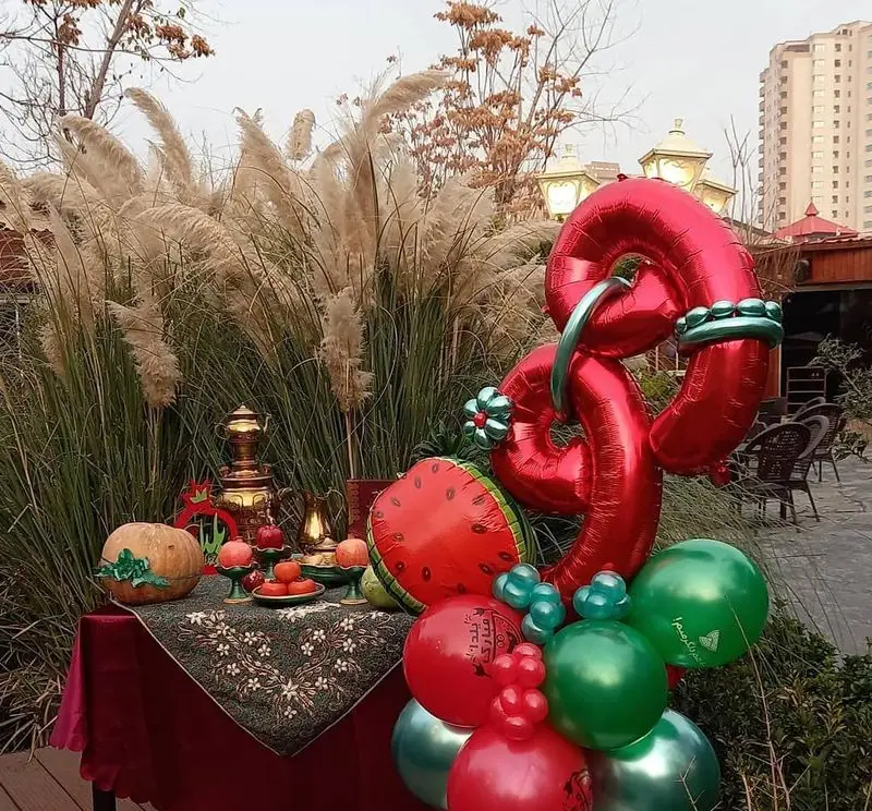 جشن شب یلدا در فضای باز رستوران و کافی شاپ دورهمی. دکور یلدایی با میز سنتی، هندوانه، انار، کدو و بادکنک‌آرایی قرمز و سبز.