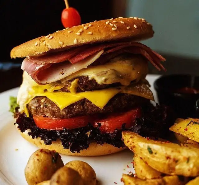 چیزبرگر دوبل لذیذ رستوران دورهمی شامل دو لایه گوشت، پنیر، بیکن، گوجه، کاهو و نان کنجدی، سرو شده با سیب زمینی سرخ شده.