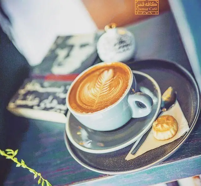 فنجان لاته آرت با طرح برگ زیبا، همراه با شیرینی کوچک و قاشق در سینی، در محیط دلنشین رستوران و کافی شاپ دورهمی.