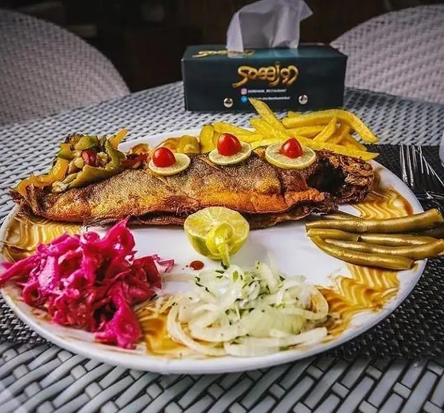 بشقاب غذای دریایی لذیذ: ماهی سرخ شده مجلسی با سیب زمینی سرخ کرده، سالاد کلم قرمز و پیاز، لیمو و مخلفات در رستوران دورهمی.