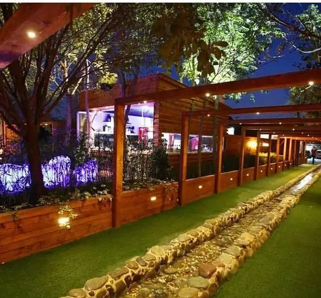 نمایی از فضای باز شبانه رستوران و کافی شاپ دورهمی با سازه‌های چوبی، مجرای سنگی آب، چمن مصنوعی و نورپردازی متنوع.