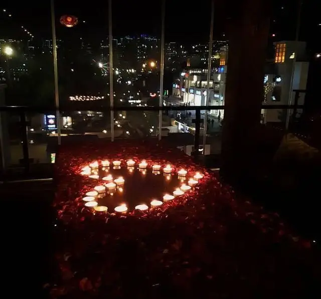 منظره شبانه زیبای شهر از رستوران و کافی شاپ دورهمی با چیدمان عاشقانه شمع و گلبرگ قرمز به شکل قلب، فضایی دلنشین برای قرار