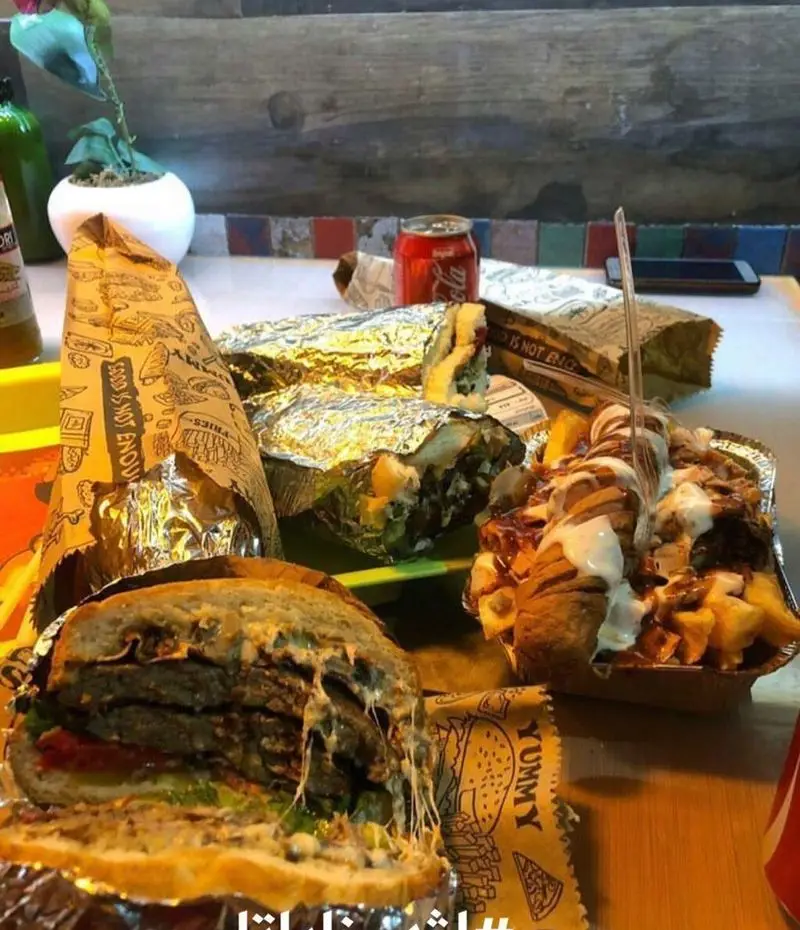 نمای نزدیک از میز شلوغ فست فود اژدر زاپاتا با همبرگر دبل پنیر آب شده، ساندویچ‌های پرملات و سیب زمینی‌های هیجان‌انگیز.