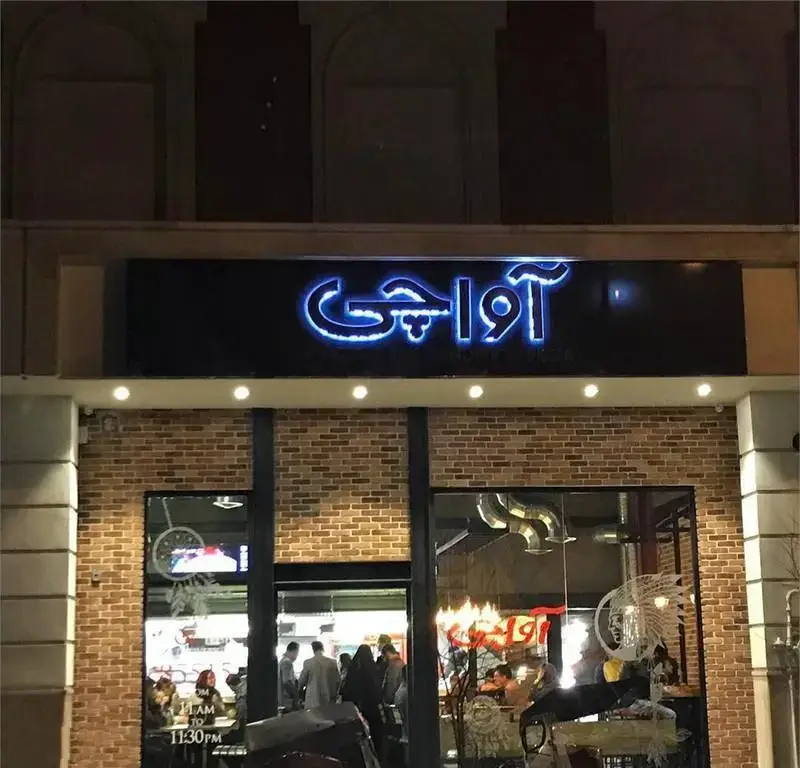 نمای بیرونی رستوران و پیتزا آپاچی در شب با تابلوی نئونی درخشان فارسی، نمای آجری زیبا و مشتریان در حال صرف غذا.