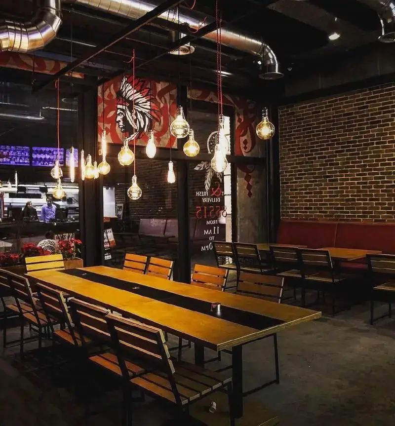 دکوراسیون داخلی مدرن و صنعتی رستوران آپاچی با میزهای چوبی بلند، نورپردازی آویز لامپ‌های ادیسون و دیوار آجری. فضایی دنج و خاص.