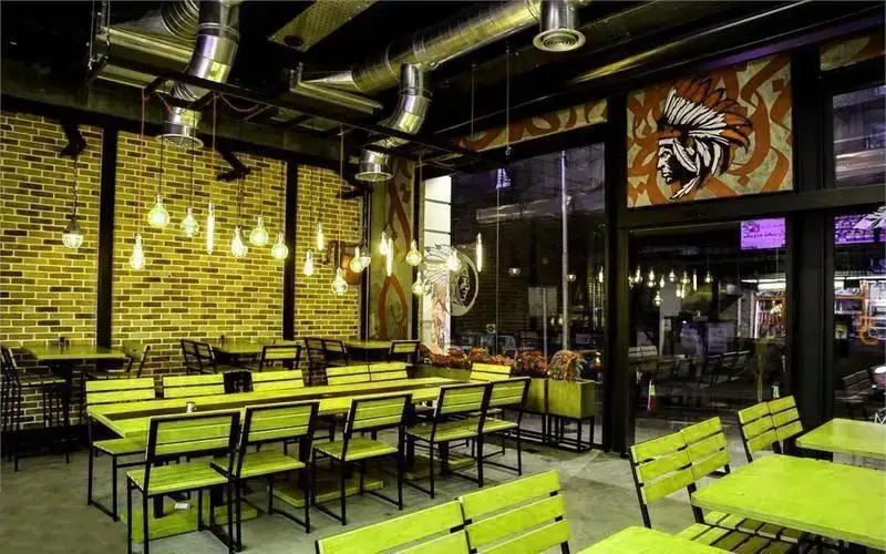 نمایی از فضای داخلی رستوران آپاچی با دکوراسیون صنعتی، شامل سقف با لوله‌های فلزی، دیوار آجری، مبلمان سبز و نماد سرخپوستی.