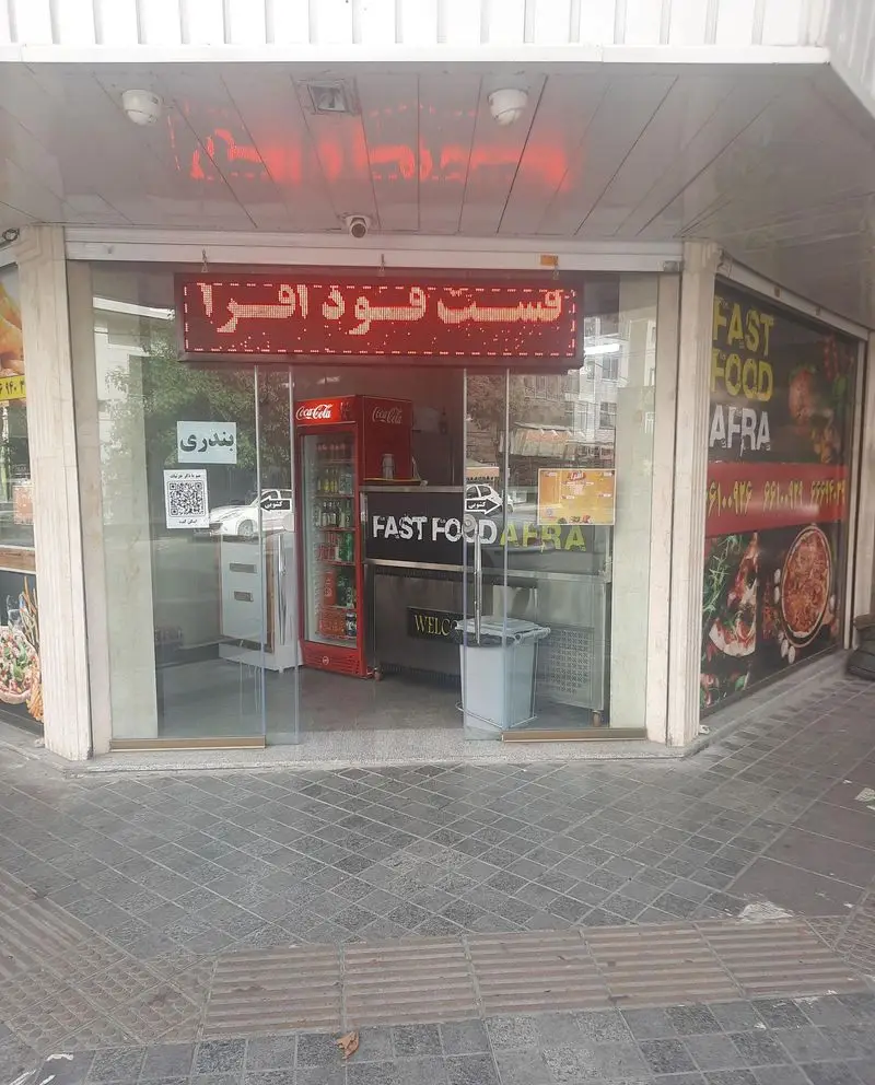 نمای بیرونی فست فود افرا با تابلوی LED قرمز و ورودی شیشه‌ای، به همراه یخچال نوشابه و تصاویر غذا در ویترین، نشان‌دهنده یک کسب و کار فعال است.