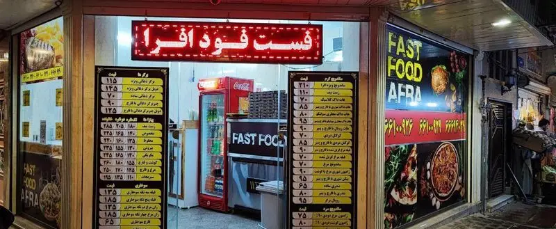 نمای شب فست فود افرا با تابلوهای روشن، منوی کامل قیمت غذاها شامل پیتزا، ساندویچ، برگر و محیط جذاب آن در شهر.