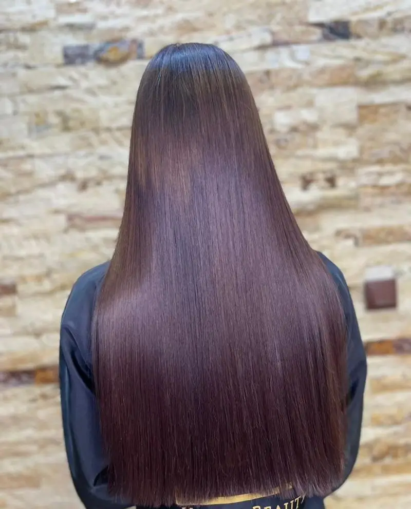 نمای پشت سر از موهای بلند، صاف و احیا شده با رنگ قهوه‌ای تیره در سالن زیبایی پانیذ خانی روی زمینه دیوار آجری روشن.