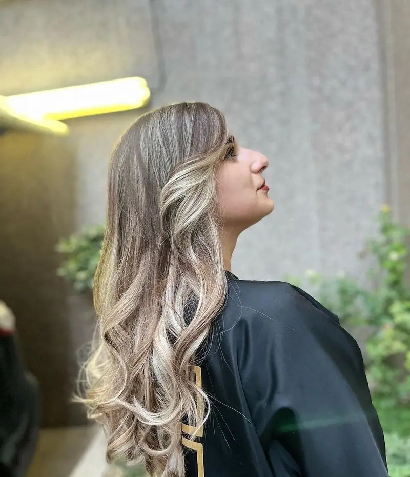 تصویر نیم‌رخ از رنگ و لایت و بالیاژ موهای بلند و موج‌دار زنانه با تناژ بلوند و قهوه‌ای در سالن زیبایی پانیذ خانی