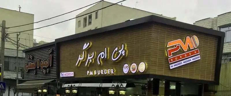 نمای بیرونی روشن و جذاب رستوران فست فود پی ام برگر با تابلوی فارسی و انگلیسی، لوگوی همبرگر و پیتزا و شماره‌های تماس برای سفارش.