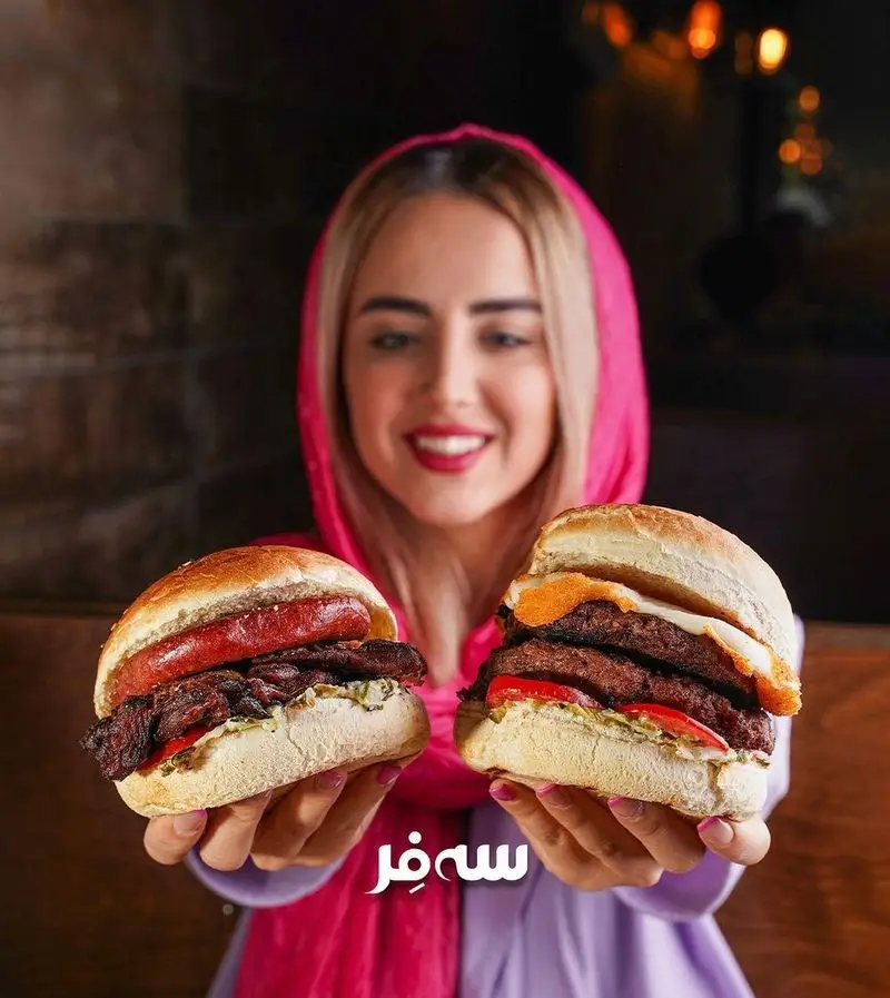 زن جوان خندان با روسری صورتی، دو برگر ویژه فست فود سه فر (یکی با سوسیس و دیگری همبرگر با تخم مرغ) را به نمایش می‌گذارد.