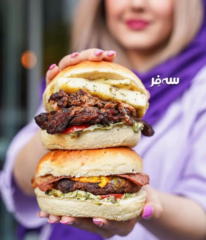 دو برگر خوشرنگ و بزرگ (مرغ گریل و گوشت با پنیر) از فست فود سه فر در دست زنی با لبخند و ناخن‌های صورتی. غذایی لذیذ و پرطرفدار.