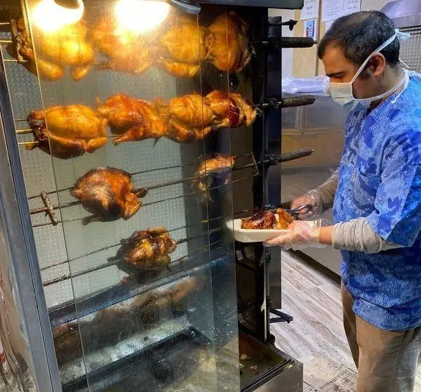 سرآشپز مرغ بریان ملاصدرا با رعایت بهداشت، ماسک و دستکش، مرغ های طلایی بریان شده را از دستگاه روتیسری سرو می کند.