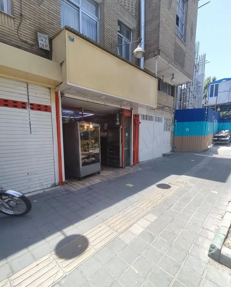 تصویری از نمای جلویی فروشگاه مرغ بریان ملاصدرا با ویترین داخلی غذا، درب ورودی باز، پیاده‌رو و ساختمان‌های آجری اطراف در روز روشن.