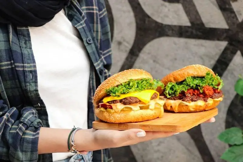 دو همبرگر خوشمزه و لذیذ فست فود شیلا، شامل یک چیزبرگر با پنیر و کاهوی تازه، روی تخته چوبی، آماده سفارش آنلاین از منوی شیلا.