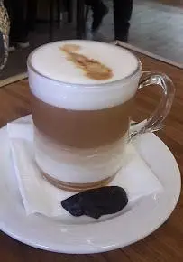 لاته لایه ای و خوش طعم کافه قنادی اوریانت با فوم شیر و تزئین قهوه روی آن، سرو شده در لیوان شیشه‌ای همراه با شکلات.