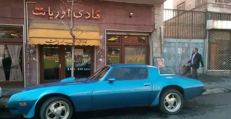 نمای خارجی کافه قنادی اوریانت با تابلوی نئونی و آفتابگیر زرد، شامل ماشین کلاسیک آبی رنگ و مردی در حال عبور در خیابان.