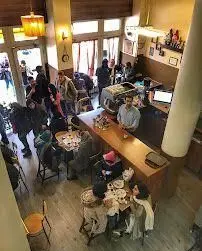 کافه قنادی اوریانت: نمای داخلی گرم و پر جنب و جوش کافه با مشتریانی که از قهوه و شیرینی لذت می‌برند. پیشخوان بلند و دکوراسیون دلنشین.