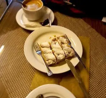 لذت دسر شیرین و قهوه تازه در کافه قنادی اوریانت: سه برش اشترودل پودرپاشیده، به همراه یک فنجان کاپوچینو لذیذ روی میز کافه.