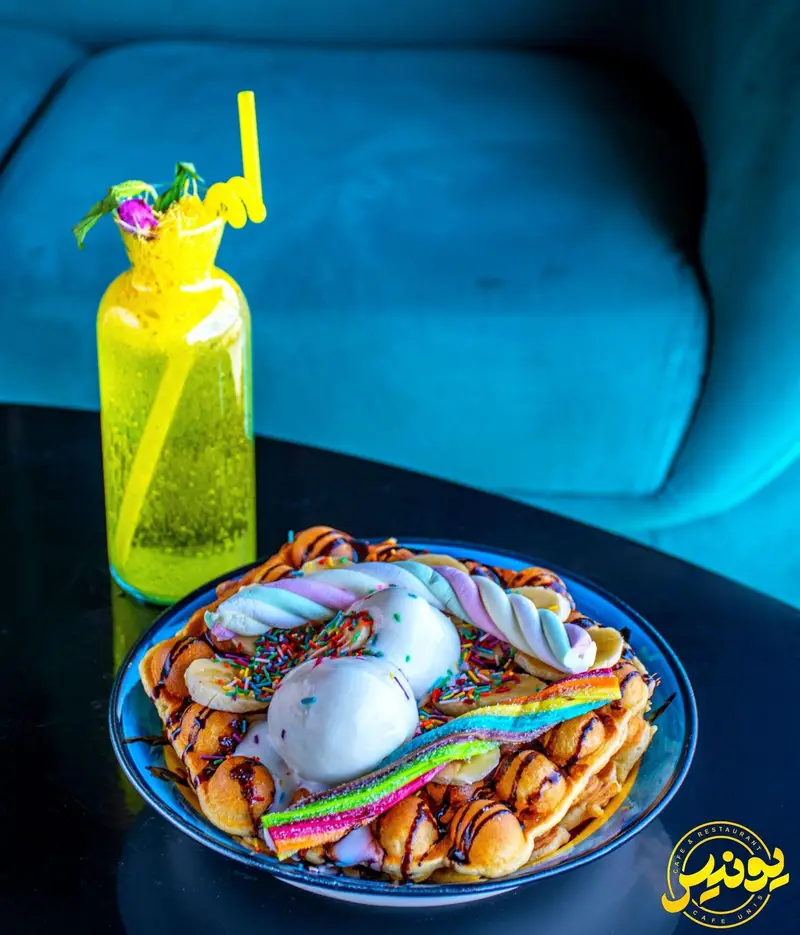تصویری دلنشین از وافل هنگ کنگی ویژه کافه یونیس با بستنی، مارشمالو، موز، اسمارتیز و سس شکلات، در کنار نوشیدنی زرد و شاداب.