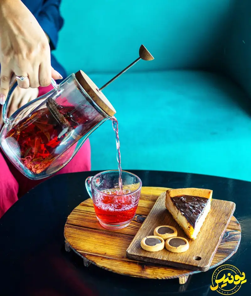 سرو دمنوش قرمز و دلپذیر همراه با چیزکیک شکلاتی و کوکی‌های خوشمزه در فضایی گرم و دلنشین کافه و رستوران یونیس.