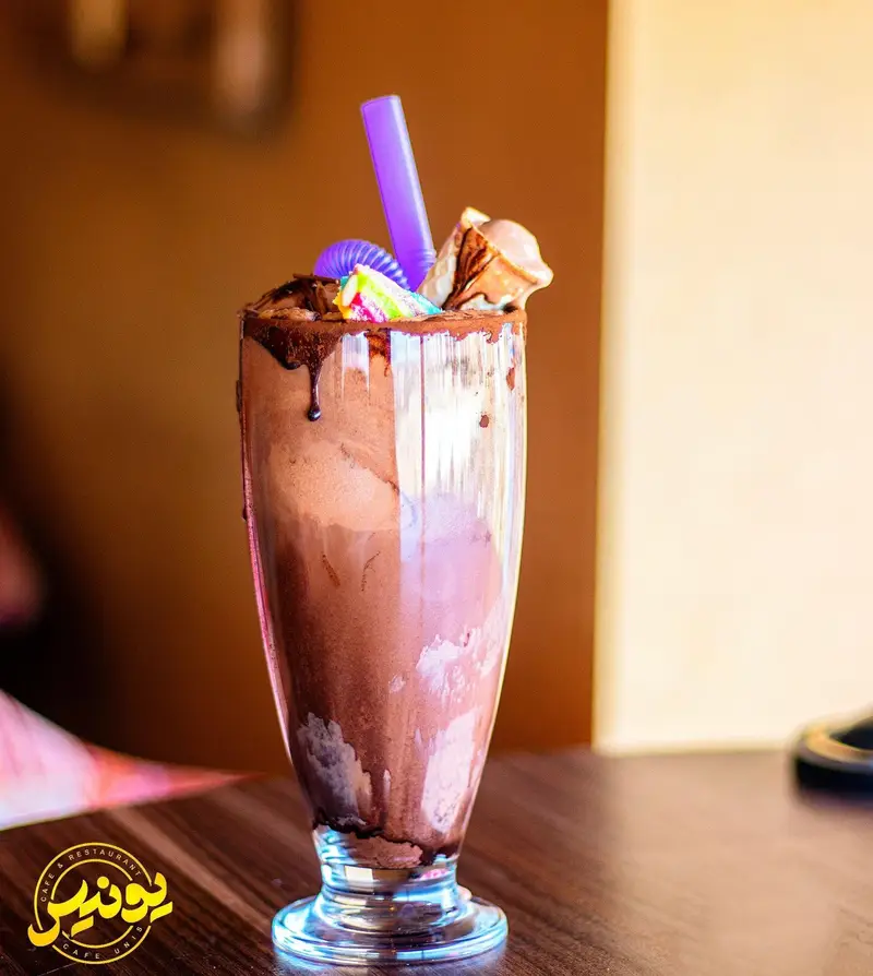 میلکشیک شکلاتی غلیظ و خوشمزه کافه یونیس، تزیین شده با سس شکلات، نی بنفش و مارشمالو رنگی، نوشیدنی دلنشین و عالی در کافه.