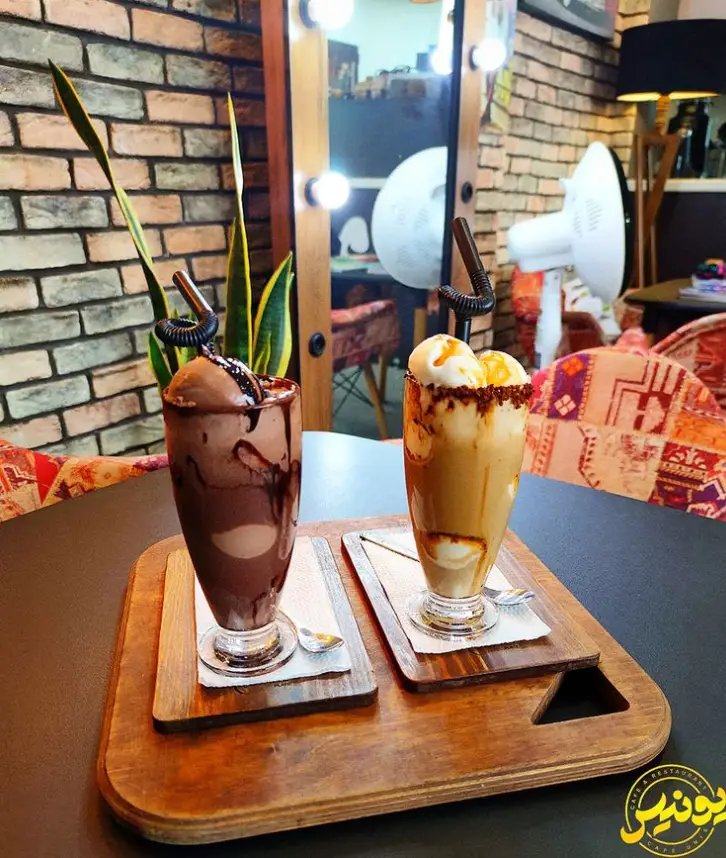نوشیدنی‌های شیک شکلاتی و بستنی وانیلی کافه یونیس، با سس کارامل و شکلات، روی سینی چوبی در محیطی با دیوار آجری و آینه نورانی.