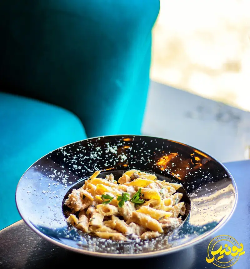 تصویری جذاب از پاستا پنه کرم‌دار در کافه یونیس، غذایی لذیذ و دلنشین در بشقابی شیک با تزیین پنیر. مناسب برای تجربه‌ای به یادماندنی.