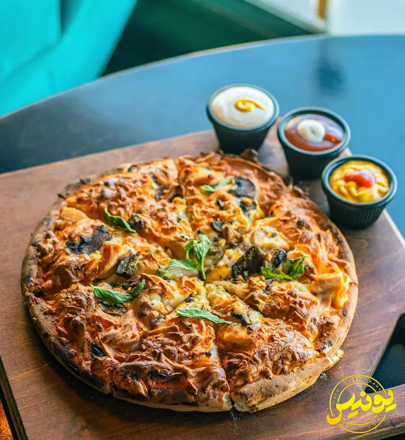 پیتزا ویژه و اشتهاآور کافه یونیس، با قارچ و پنیر طلایی، سرو شده روی تخته چوبی همراه با سس‌های متنوع. تجربه‌ای لذیذ از منو.