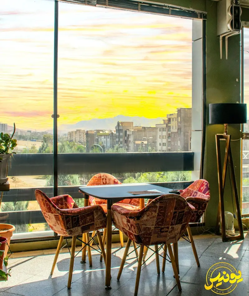 کافه یونیس: فضایی دنج با میز و صندلی‌های طرح‌دار، پنجره‌ای بزرگ رو به منظره زیبای غروب آفتاب و شهر، ایده‌آل برای لذت بردن از لحظات.