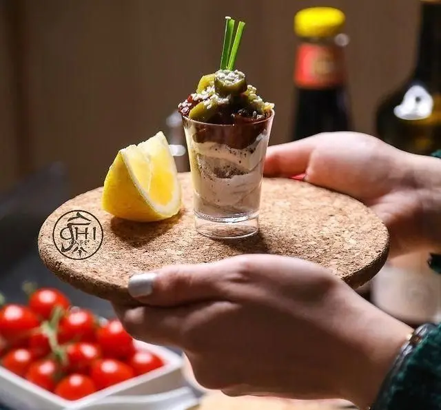 دسر/مزه لایه‌ای خاص و خوش‌طعم چی کافه، تزئین شده با سبزیجات تازه و لیمو، روی سینی چوب پنبه‌ای با لوگوی چی کافه.