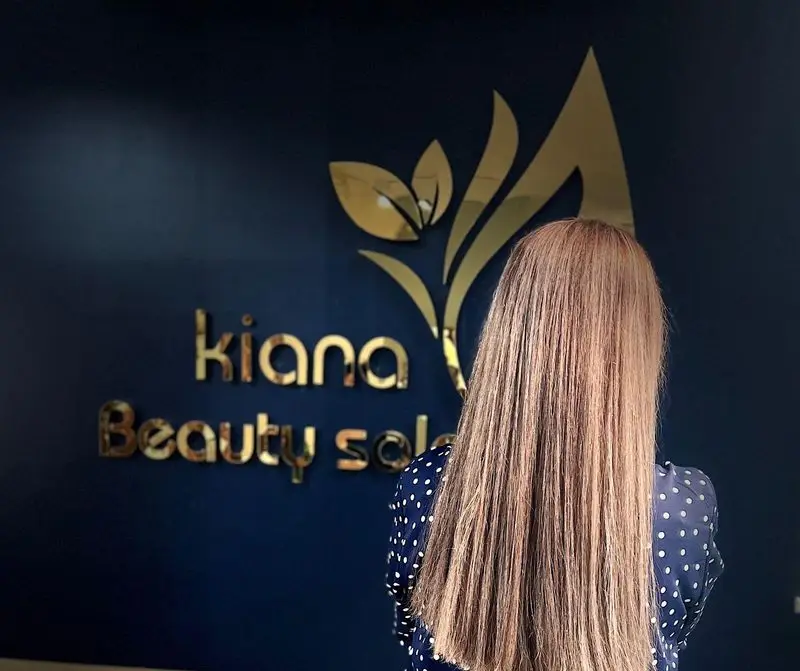 مشتری با موهای بلند و خوش‌حالت در سالن زیبایی کیانا، دیوار آبی با لوگوی طلایی Kiana Beauty Salon و اسم برند برای خدمات مو و زیبایی.