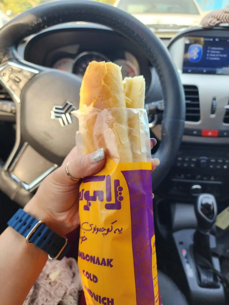 ساندویچ ژامبونک داخل ماشین: مشتری ساندویچ سرد بسته بندی شده فست فود ژامبونک را در دست گرفته، لوگو سایپا روی فرمان خودرو مشخص است.
