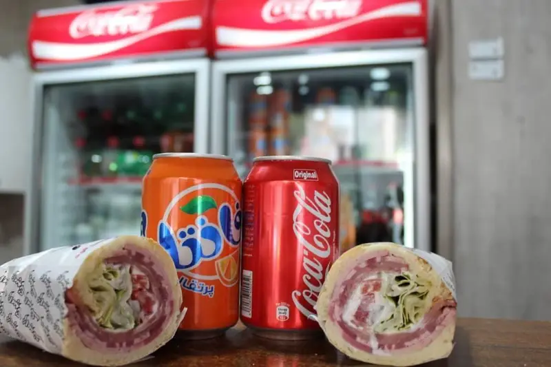 ساندویچ‌های رول‌شده و تازه فست فود ژامبونک در کنار نوشابه‌های کوکاکولا و فانتا، گزینه‌ای عالی برای یک وعده کامل.