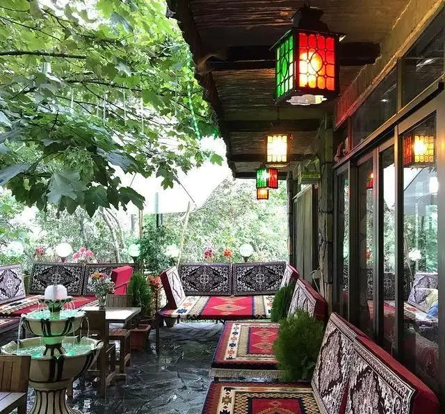 فضای سرسبز و دلنشین رستوران جویبار، با تخت‌های سنتی، فرش‌های زیبا، آبنما و فانوس‌های رنگی زیر سایه درختان.
